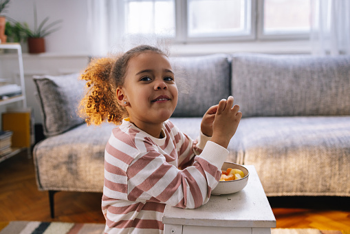 Portrait of a young Afro-American girl eating fruit from the bowl, looking at camera, smiling.