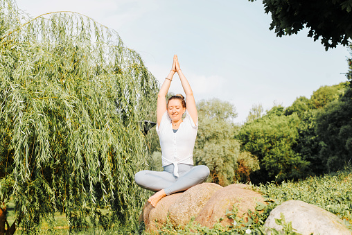Happy young woman with closed eyes meditating in nature, practicing yoga asana, sitting on a stone outdoors. Yogi woman enjoying morning workout in the park. Yoga and meditation, healthy lifestyle.