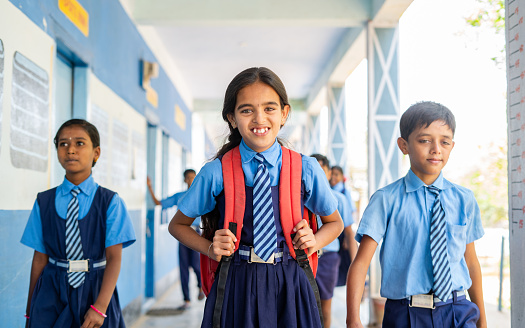 Happy girl kid in uniform standing at school corridor while other kids passing - concept of confidence, education, childhood growth and development
