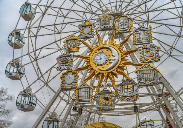 Ferris wheel with a musical clock in the form of a sun disk and the moon, surrounded by opening windows. Ferris wheel with a musical clock in the form of a sun disk and the moon, surrounded by opening windows. close-up from below. Tyumen amusement park (Siberia, Russia). Gray cloudy sky on an autumn day western siberia stock pictures, royalty-free photos & images