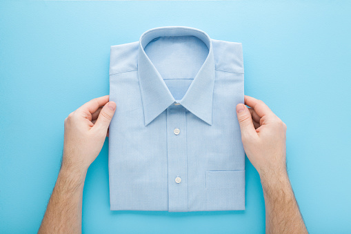 Young adult man hands holding folded classic shirt on light blue table background. Pastel color. Point of view shot. Closeup. Male clothes. Top down view.