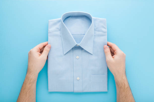 Young adult man hands holding folded classic shirt on light blue table background. Pastel color. Point of view shot. Closeup. Male clothes. Top down view. stock photo
