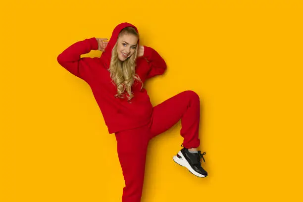Smiling woman is posing on one led in red sweatpants and hooded sweatshirt.