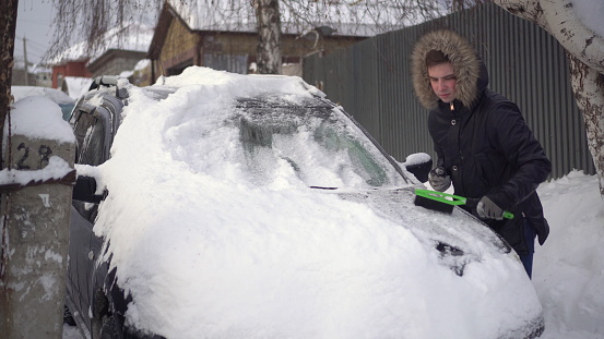 A young man in a jacket cleans his car of snow with a brush. The car was covered in snow. Weather disaster. 4k