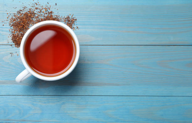 Ceramic cup of aromatic rooibos tea and scattered dry leaves on light blue wooden table, flat lay. Space for text