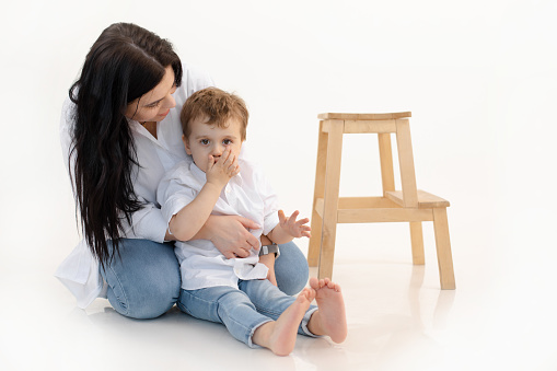 Caring woman and boy hugging and spending time together. Close bounding between mother and little son, quieting, sitting on the floor. Wooden chair on white background. Studio shot. Family weekend