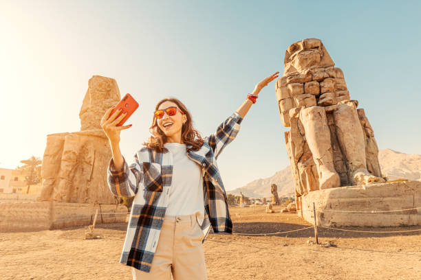 Travel blogger girl takes selfie pictures on a smartphone at the famous two Colossi of Memnon - massive ruined statues of the Pharaoh Amenhotep III. World tourism attractions stock photo