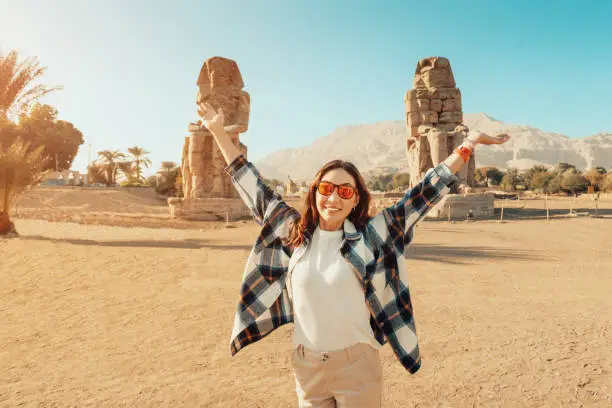 Happy tourist girl interested in Egyptology and archaeology and gets a travel experience at the Memnon colossus Amenhotep pharaoh statue in Luxor.