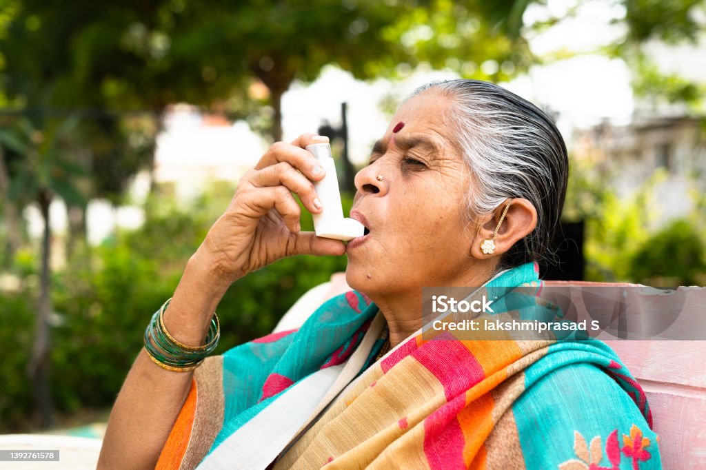 elderly woman using asthma inhaler while sitting at park due to allergy - concept showing effects of pollution, illnes and disease elderly woman using asthma inhaler while sitting at park due to allergy - concept showing effects of pollution, illness and disease. Asthma Inhaler Stock Photo