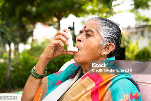 Elderly Woman Using Asthma Inhaler While Sitting At Park Due To Allergy Concept Showing Effects Of Pollution Illnes And Disease Stock Photo - Download Image Now