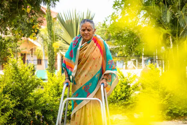 Photo of old woman walking with support of walker due to knee joint pain at park - concept of arthritis, support and rehabilitation