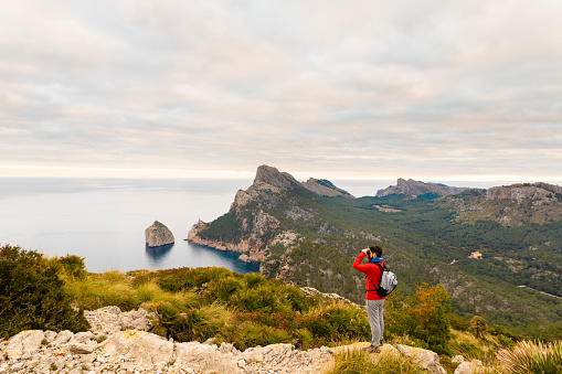 Hiker standing on a rock looking into the distance with binoculars in front of an amazing landscape on the north coast of Mallorca. Color editing. Part of a series.