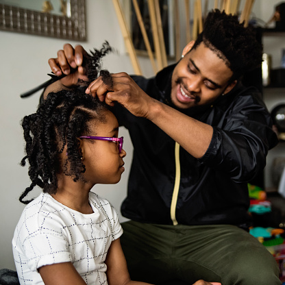 Father undoing braids of young daughter in living room. Dad is in thirties, daughter is 7 year’s old. Square waist up indoors shot in natural light. Some copy space.