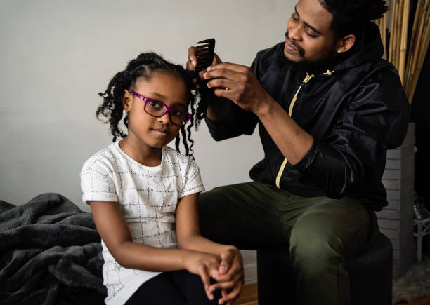 Father undoing braids of young daughter in living room. stock photo