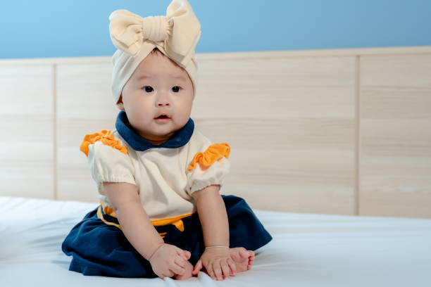 adolable Asian newborn baby girl, big eyes, puffy cheeks, cutely dressed in yellow and blue, sitting on a white bed looking at something, with a happy smiling face. adolable Asian newborn baby girl, big eyes, puffy cheeks, cutely dressed in yellow and blue, sitting on a white bed looking at something, with a happy smiling face. cutely stock pictures, royalty-free photos & images