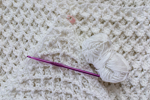 Top view of on a partially finished white cotton blanket, original raised crochet stitch pattern, a ball of yarn, 5 mm purple crochet hook, yellow measuring tape, pink marker. Handmade creativity