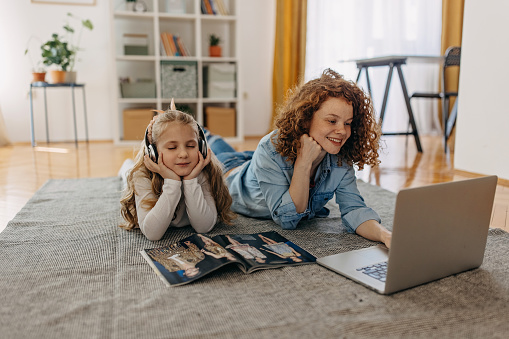 Mother is one the floor with her daughter. Mother is using the laptop and daughter is wearing wireless headphone and listing the music