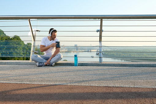 Middle aged sportsman, athlete in headphones sits on a city bridge and checks a mobile application with monitoring heart rate and calories burned during a workout on his phone in a smartphone holder