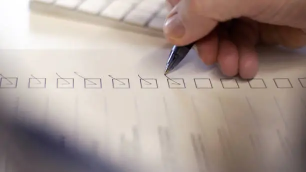 Close up image of a person’s hand using a ballpoint pen to put a check or tick mark in a row of boxes on a paper form sheet. In an office environment.