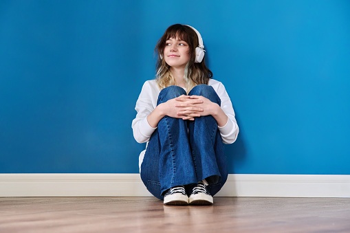 Portrait of a teenage girl in headphones sitting on the floor on a blue background. Beauty, youth, fashion, color, young people concept