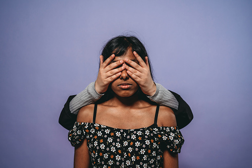 Conceptual shot of a woman with eyes covered by another woman's hands. Purple background.