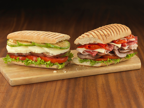 Two Italian sandwich (panini). One of them stuffed with tomato, lettuce, eggplant, mozzarella cheese and zucchini. The other stuffed with lettuce, tomato, smoked turkey, ham, salami, gouda cheese and roasted red bell peppers.