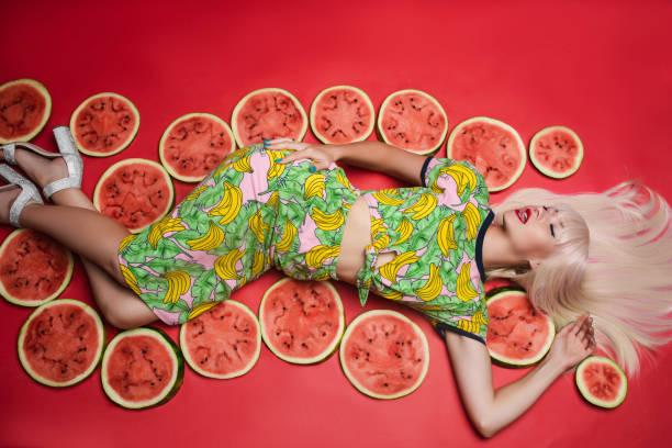 Sexy beautiful young woman lying on floor posing for photo shooting surrounded by watermelon Sexy beautiful young woman lying on floor posing for photo shooting surrounded by watermelon full shot. Charming blonde female model in colorful clothing relaxing isolated at red studio background seduction stock pictures, royalty-free photos & images