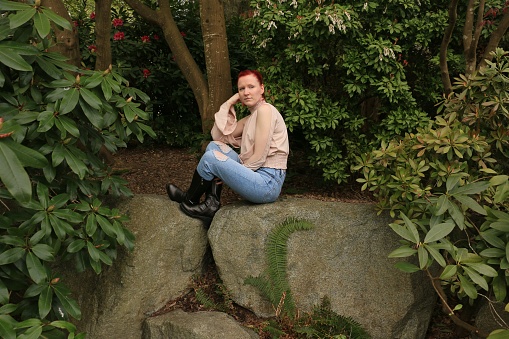 A Caucasian woman sitting on a rock hidden in a garden. She is wearing a beige top, blue jeans that are torn and black knee length boots.