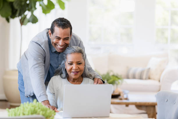 el esposo mayor mira por encima del hombro de la esposa a la pantalla de la computadora portátil - home finances couple computer african ethnicity fotografías e imágenes de stock