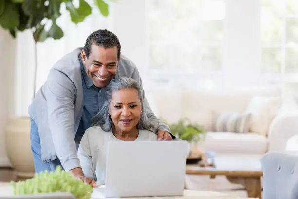 Photo of Senior husband looks over wife's shoulder at laptop screen