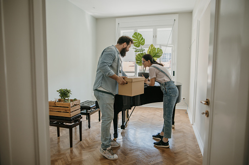 Couple in their new apartment, they are unpacking in their ne home together. Woman is putting potted plant on a piano.