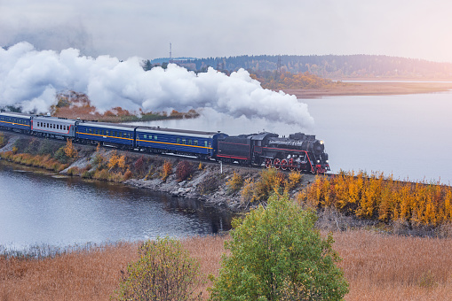 Retro steam train moves along the lake at autumn cloudy morning.