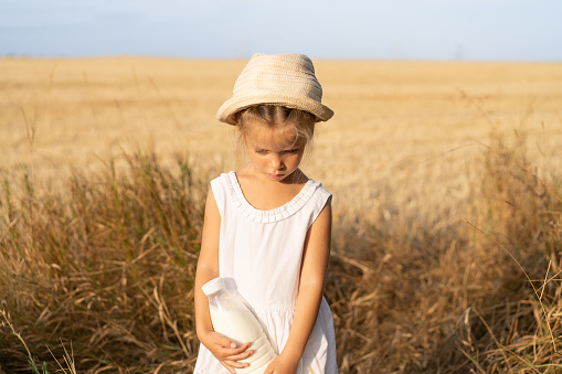 Little girl standing wheat field holding bottle of milk in hand dressed white dress and straw hat. Healthy eating concept, rural scene. Caucasian female child with milk outdoor. Blue sky background