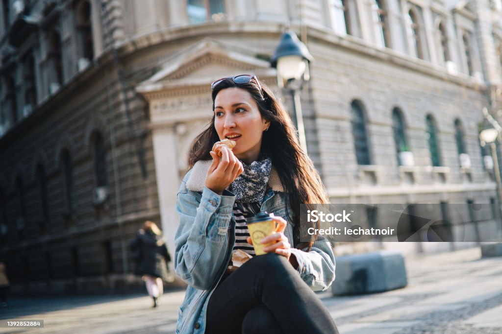 woman having a coffee break in the city Young woman still wearing a fashionable bandana after the pandemic is over, having a coffee and a croissant break while checking on the apps on her mobile phone. Coffee - Drink Stock Photo