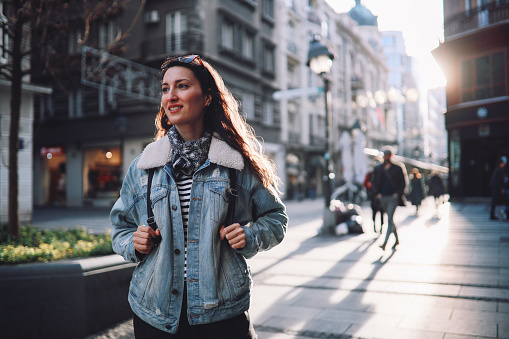 Young woman in her late 30s enjoying a quick city break, walking down the city centre. She is casually dressed, wearing a denim jacket.