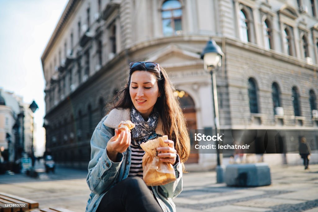 young woman eating a croissant on the street Young woman on a city break after the pandemics, walking in downtown area, having a coffee and a croissant break. Adult Stock Photo