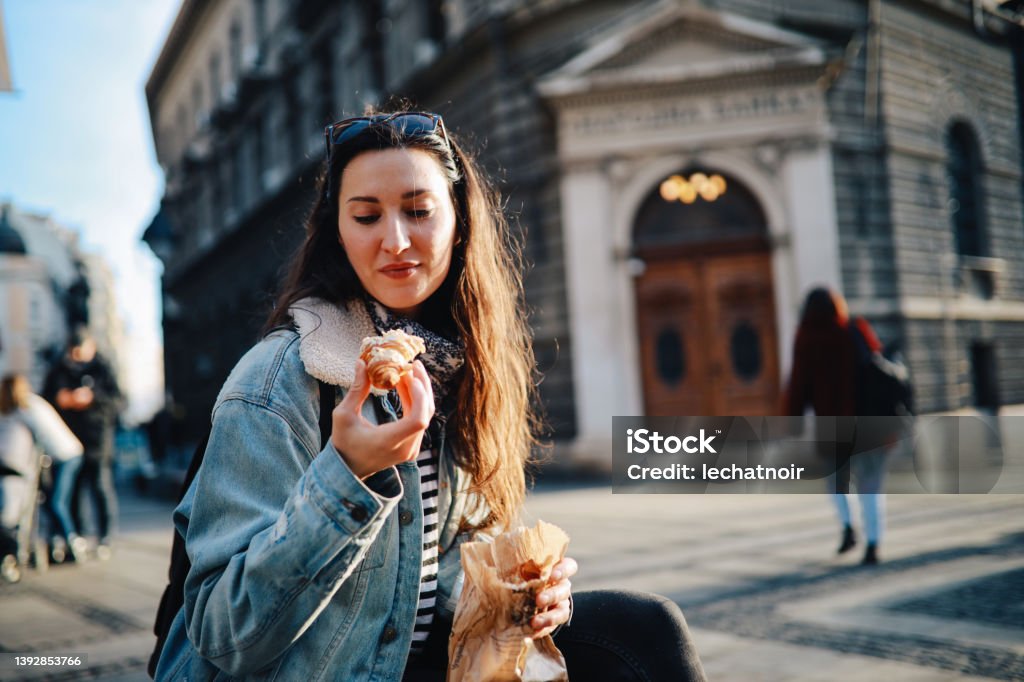 young woman eating a croissant on the street Young woman on a city break after the pandemics, walking in downtown area, having a coffee and a croissant break. Croissant Stock Photo