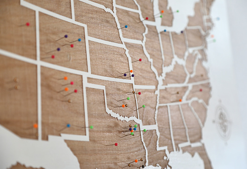 Wooden US States map on the wall with multicolored pins on it.