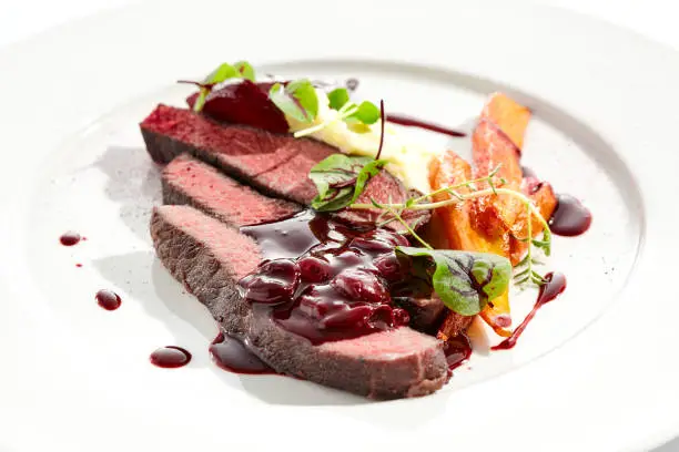 Photo of Venison steak with baked vegetables isolated on white plate. Meat steak medium rare roasted with carrot, beetroot and mashed potatoes with cherry sauce. Wild meat in restaurant menu.