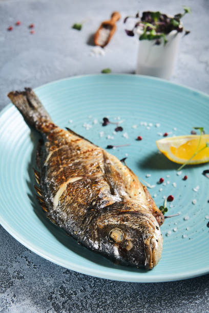 istock Grilled dorada in blue plate on stone background with textile. Roast fish in restaurant menu concept. Roasted whole dorado with lemon. Contemporary seafood menu. Mediterranean food. 1392862853