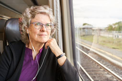 Smiling mature woman wearing earphones and looking out through a train window at the passing scenery