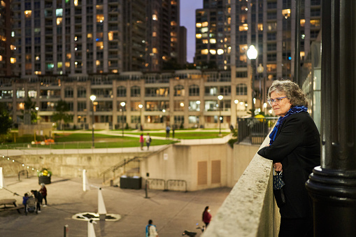 Mature woman smiling while leaning on a wall and looking out over a busy city square in the evening