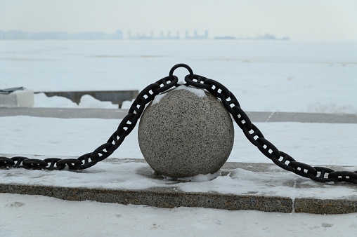 Granite ball with chains on the winter snow-covered city embankment. In the background, the silhouettes of urban buildings.