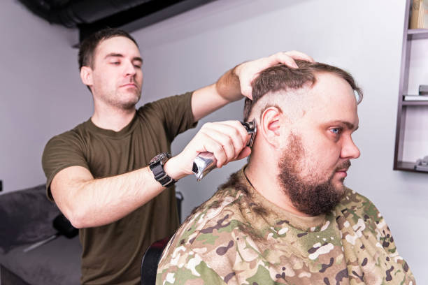 un jeune homme en uniforme militaire se rase la tête chauve pour le service militaire. un gars avec une barbe se fait couper les cheveux dans un salon de coiffure. rasez votre tête chauve - employee barracks photos et images de collection