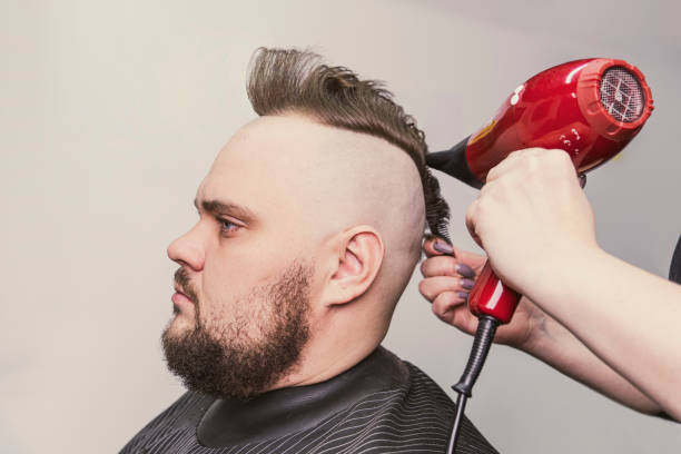 女性の理髪師は、ひげを生やした大人の男性にモホークの髪型を作ります。ヘアドライヤー付きのヘアスタイリング。 - frizzy human hair hairdresser hair dryer ストックフォトと画像