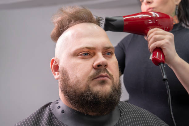 理髪店の椅子に座りながら美容師に散髪されるモホークのあごひげを生やした男。美容師の女の子が男にモホークを作ります。 - frizzy human hair hairdresser hair dryer ストックフォトと画像