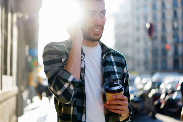 istock Young bearded hipster guy having mobile conversation with friends carefully listening while strolling around sunny town.Good looking tourist communicating via smartphone using roaming service 1392882346