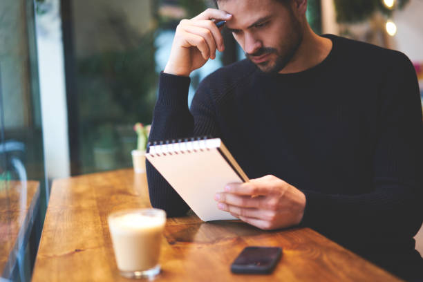 istock Cropped image thoughtful bearded young businessman creates startup project idea using notebook while working in contemporary coworking interior. Pensive student  studying in coffee shop indoors 1392882372