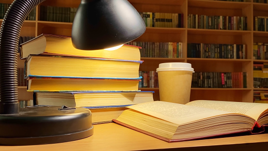 Books, coffee and lamp rotating on desk in library, book shelves in dark room