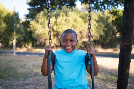 A portrait of a young African boy looking at the camera with a wide smile, he is swinging on the jungle gym at the play park